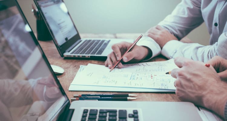 Twee personen bekijken documenten en maken aantekeningen aan een bureau met twee open laptops.