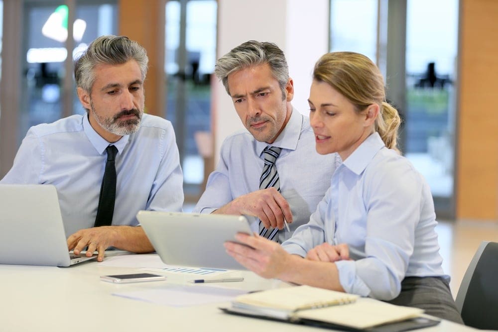 Three business professionals, two men and one woman, are seated at a table looking at a tablet. They are dressed in formal attire and appear to be discussing something on the screen.