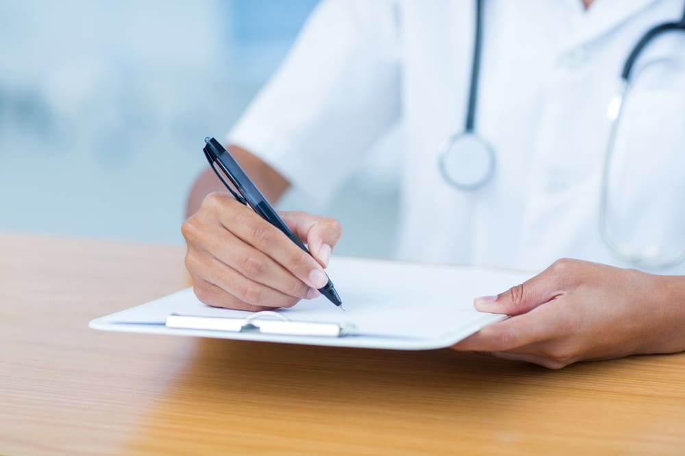 A person wearing a white coat and stethoscope writes on a clipboard at a wooden desk.