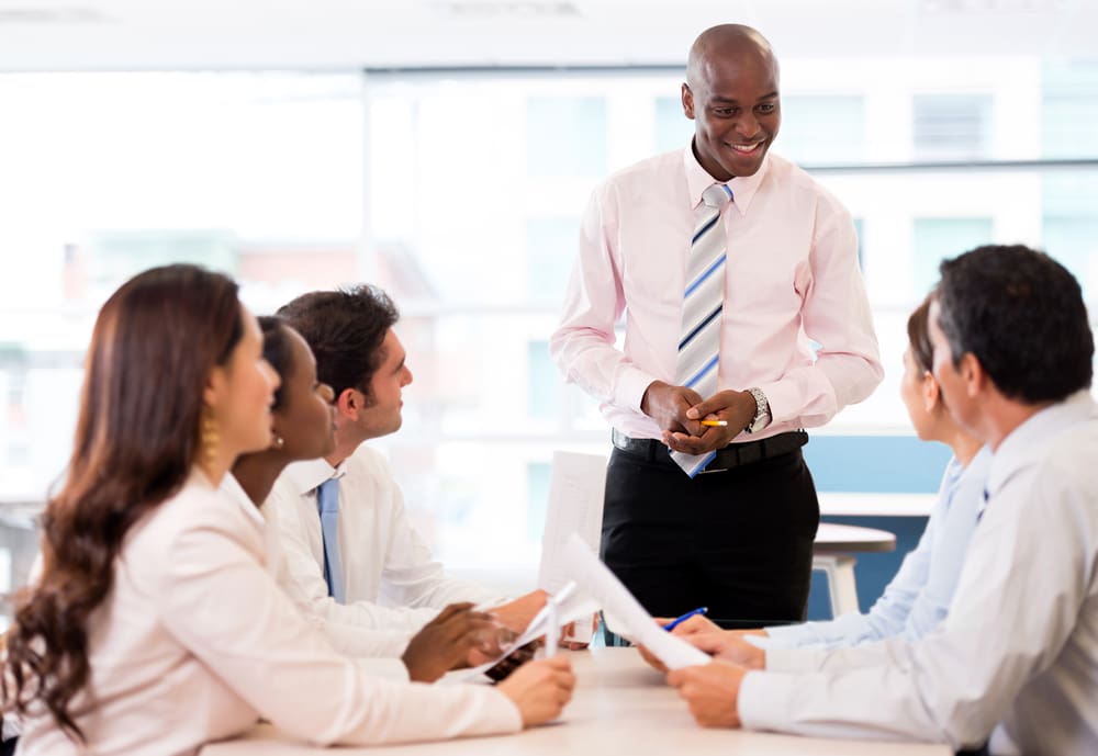 A man in a white shirt and blue-striped tie stands and smiles while addressing a group of seated colleagues in a brightly lit office.