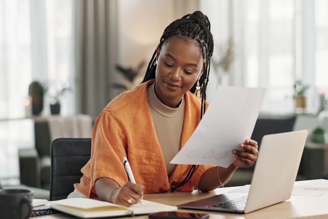 Black woman in home office, documents and laptop for research in remote work, ideas and thinking. Happy girl at desk with computer, writing notes and online search in house for freelance networking.